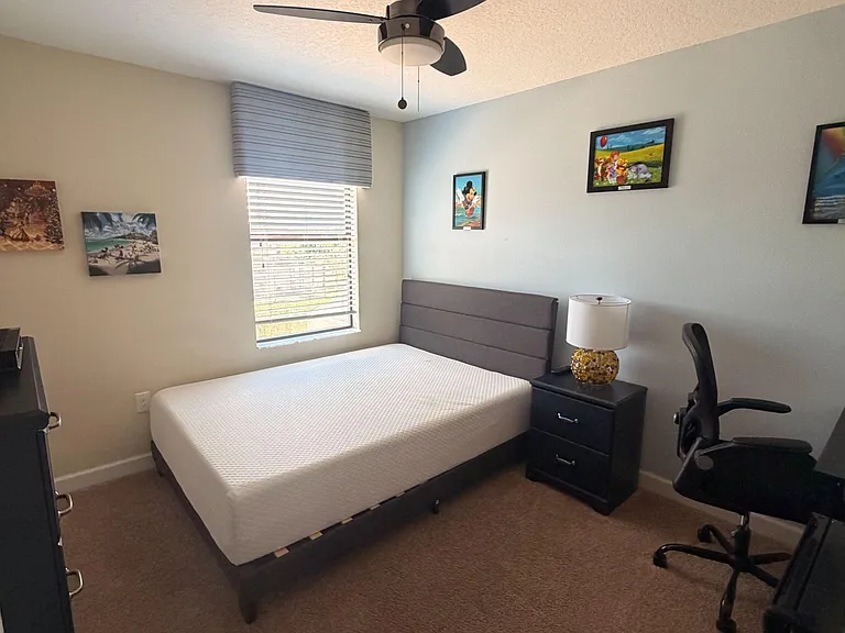 Furnished bedroom with ceiling fan, window blinds, nightstand, and desk areaideal for sleeping and studying.