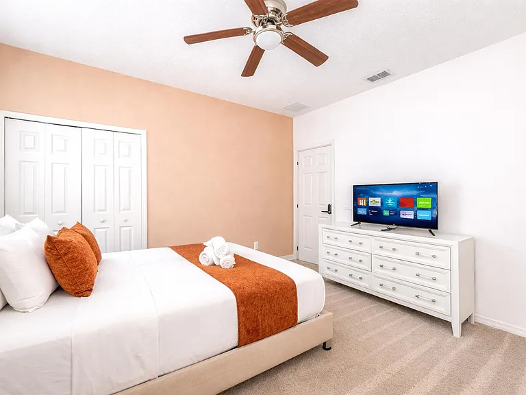The room features a stylish upholstered bed, neutral carpeting, a ceiling fan, and ample natural light.