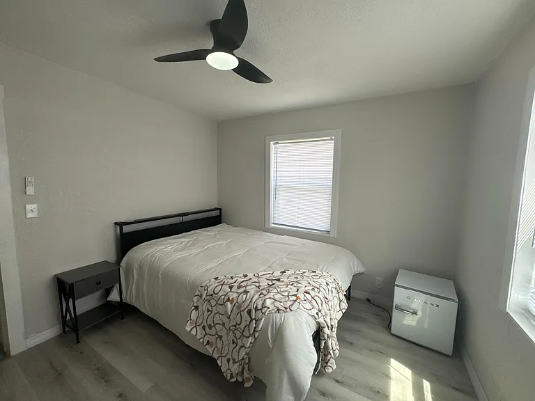 Private bedroom with a ceiling fan, bedside table, and window with blinds, offering comfortable space for rest and storage.