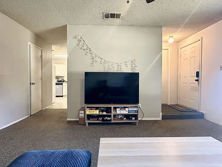 Open living area with carpeted floors, ceiling fan, and space for a media setup, connecting easily to the kitchen and entry.