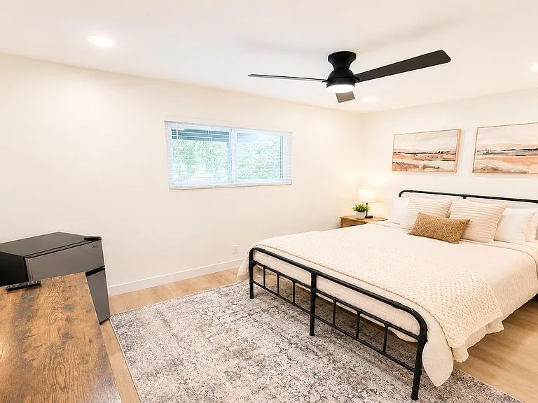 Spacious furnished bedroom with a queen bed, nightstands, ceiling fan, and a work desk plus mini fridge for convenience.