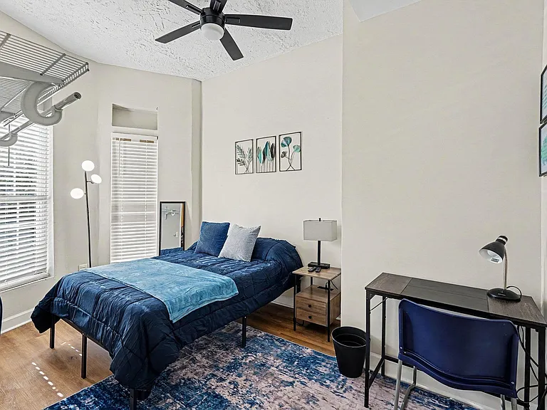 Furnished bedroom with a full bed, nightstand, and dedicated desk area, plus two windows and a ceiling fan.
