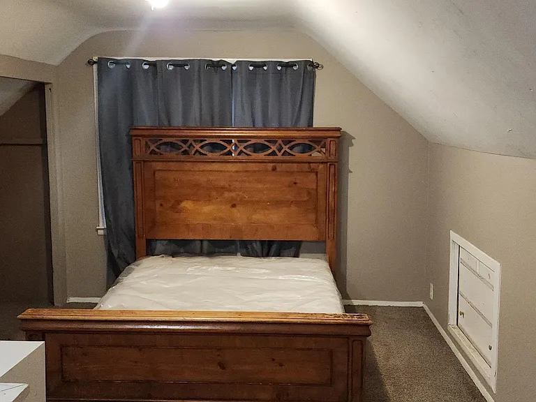 Carpeted bedroom with a full bed, sloped ceilings, and a window dressed with blackout-style curtains for privacy.