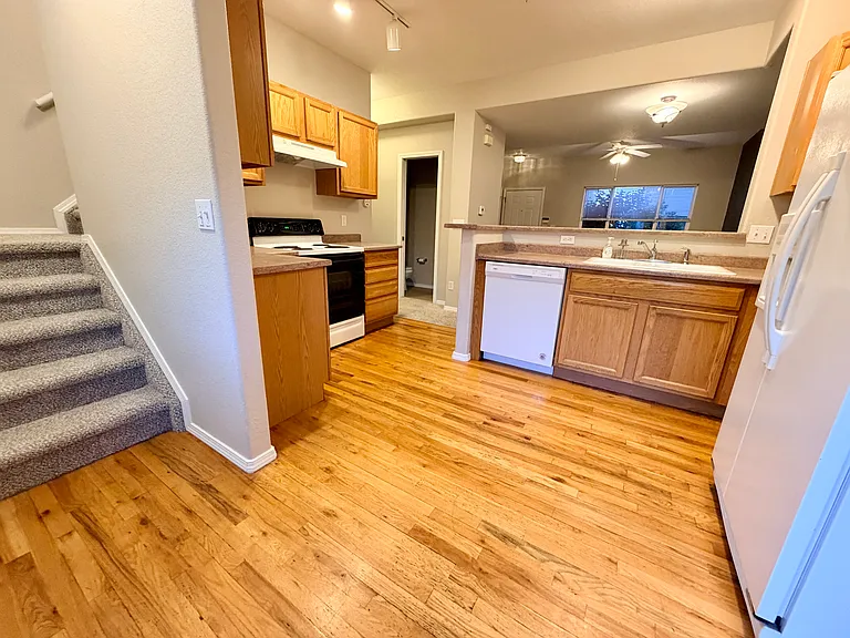 Main kitchen view showing hardwood flooring, sink, dishwasher, and open layout along with stairs that lead to the upper level. Entry is from the parking area directly in to the home.