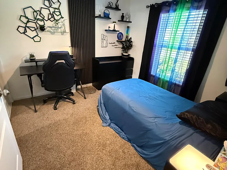 Furnished bedroom with carpet flooring, ceiling fan, dedicated desk setup, and a window with blinds for natural light.