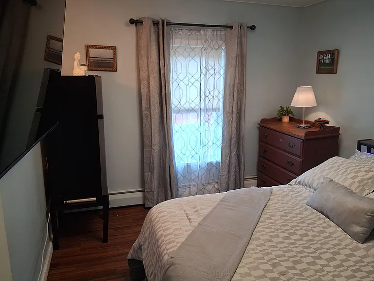 Furnished bedroom with a comfortable bed, dresser storage, and a large TV, plus a window with curtain panels for privacy.