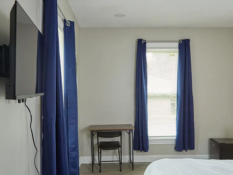 Bedroom with a large window, blackout curtains, wall-mounted TV, small desk area, and vinyl plank flooring.