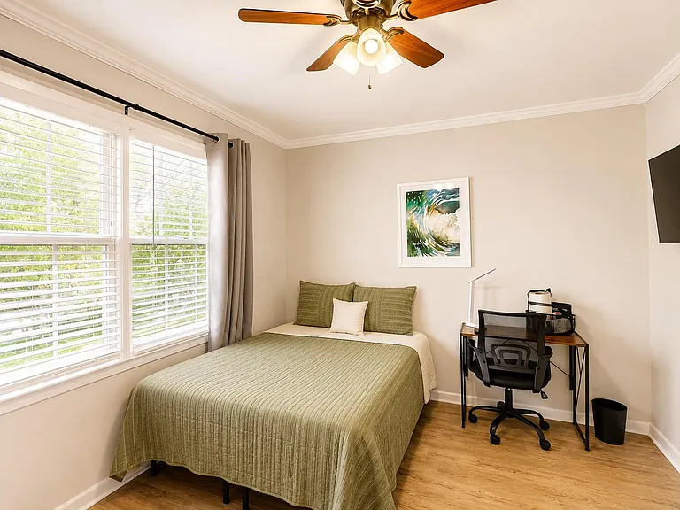 Furnished bedroom with large windows, ceiling fan, wall-mounted TV, and a dedicated desk for work or study.