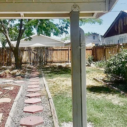 Beautifully landscaped backyard. Walkway from home to gate. Notice grass and mature rose bushes on right. Covered patio. Mature mimosa tree in background.