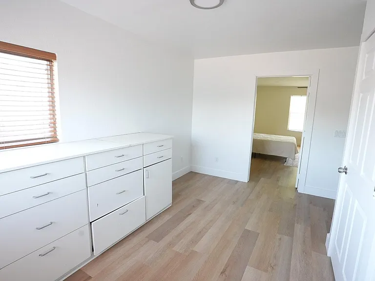Spacious room with built-in dresser storage, wide-plank flooring, and a large window with blinds for natural light.