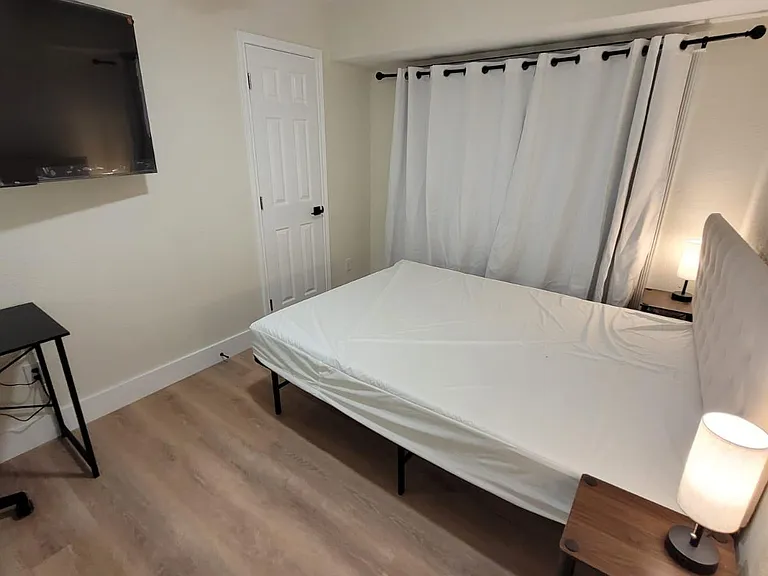 Furnished bedroom with a bed, nightstand lighting, wall-mounted TV, and ceiling fan, plus window treatments.