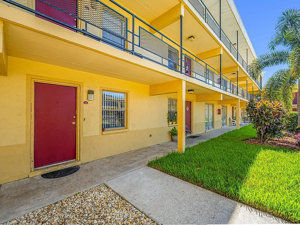 Charming apartment exterior featuring bright yellow walls, red doors, and vibrant landscaping with palm trees.