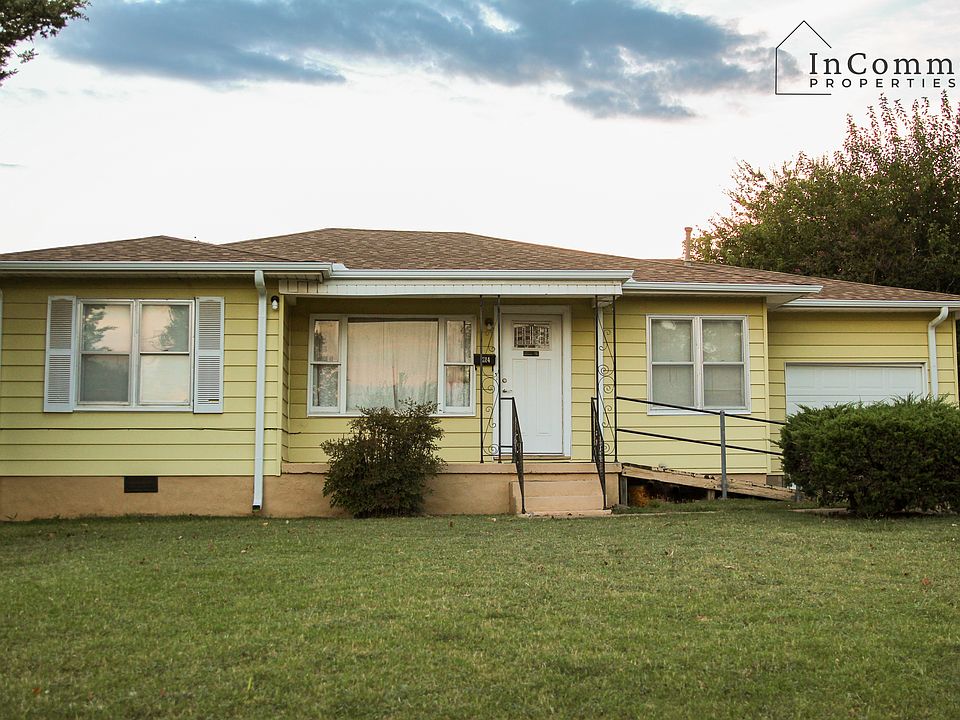 A cute single-story yellow house with white trim. It has a small covered front porch with decorative metal railings, a front-facing garage, and a ramp for accessibility.