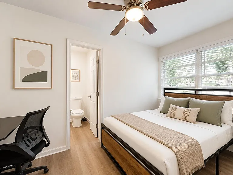 Well-appointed bedroom with a comfortable bed, work desk, ceiling fan, and large window with blinds for natural light.