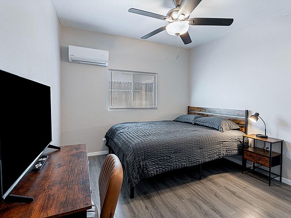 Furnished bedroom with a full bed, nightstand, ceiling fan, and window blinds plus a dedicated desk/TV setup.