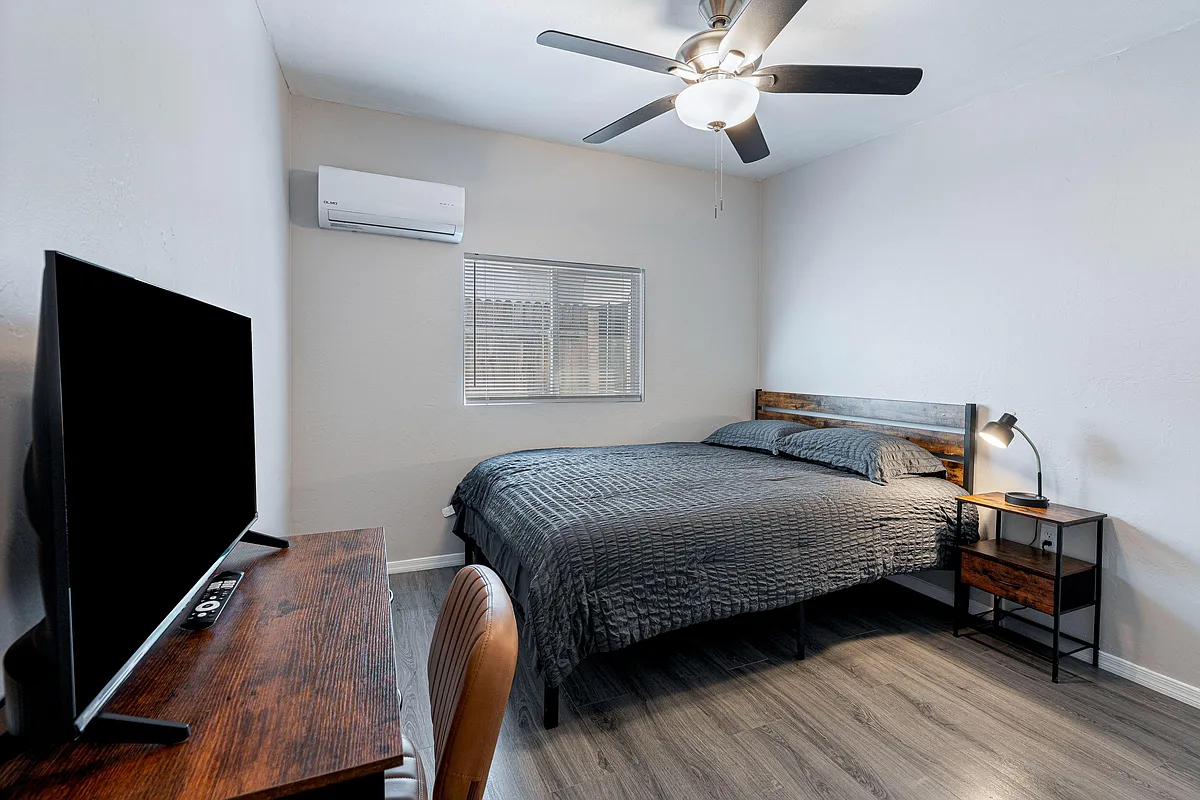 Furnished bedroom with a full bed, nightstand, ceiling fan, and window blinds plus a dedicated desk/TV setup. - E Garden St