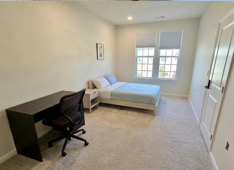 Spacious bedroom with a full bed, nightstand, and dedicated desk area, plus two large windows for natural light.