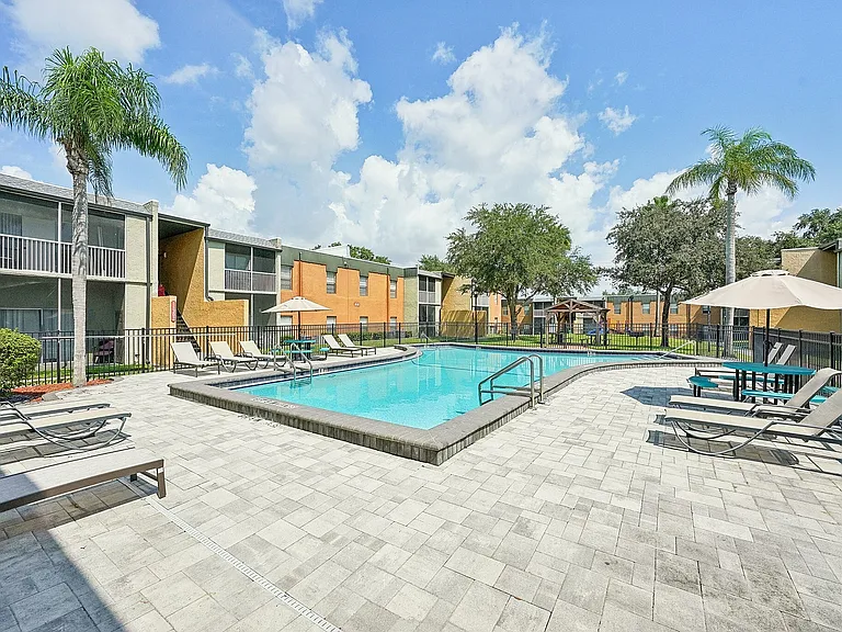 Charming apartment community with a sparkling pool, surrounded by modern lounge chairs and lush greenery under a clear blue sky.