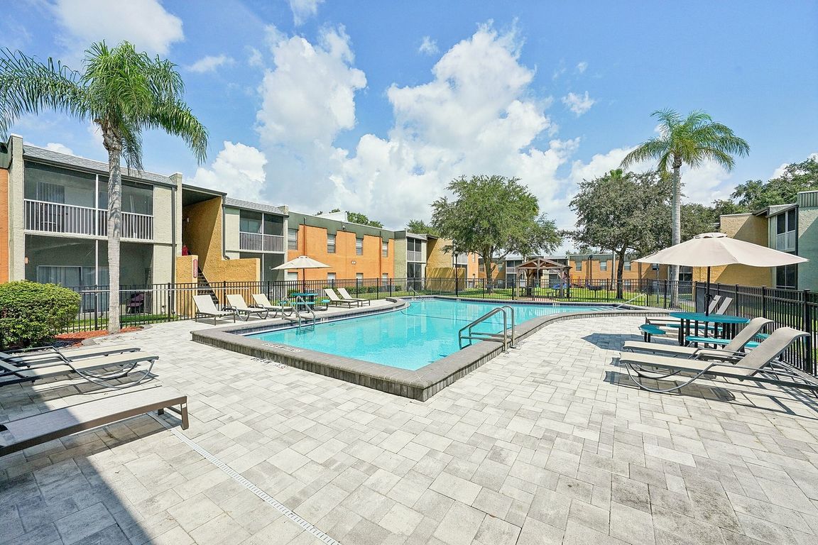 Charming apartment community with a sparkling pool, surrounded by modern lounge chairs and lush greenery under a clear blue sky.
