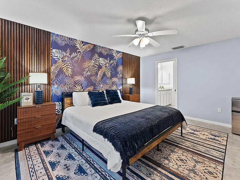 Spacious furnished bedroom with ceiling fan, nightstands, mini fridge, and direct access to an adjacent bathroom vanity.