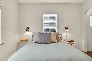 Bedroom features a comfortable layout with a centered bed, window with blinds, and convenient bedside lighting.