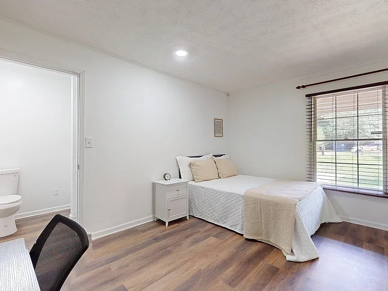 Furnished bedroom with a large window, wood-style flooring, and an en-suite bath for added privacy and convenience.