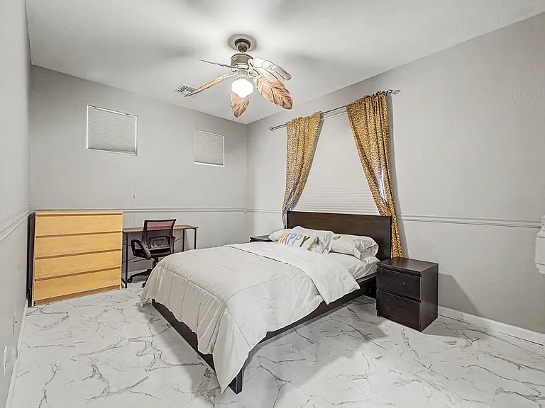 Furnished bedroom with a full bed, nightstand, dresser, and desk, plus ceiling fan lighting and window coverings.