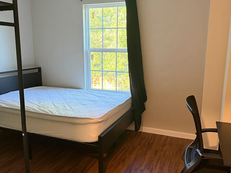 Furnished bedroom with a bed frame, desk chair, ceiling fan, and a large window with curtains for natural light.