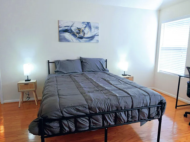 Furnished bedroom with hardwood floors, a large window with blinds, bedside tables and lamps, and a ceiling fan.