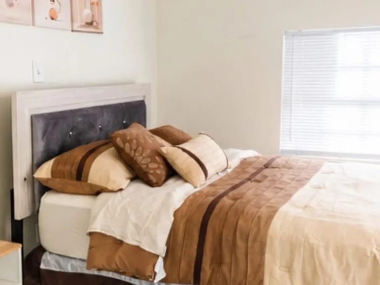Furnished bedroom with a full-size bed, upholstered headboard, and a window with blinds for light and privacy.