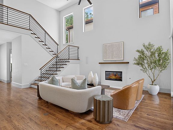 Two-story living area with high ceilings, wood-finish flooring, a contemporary fireplace, and a modern staircase featuring horizontal metal railings and wood handrails
