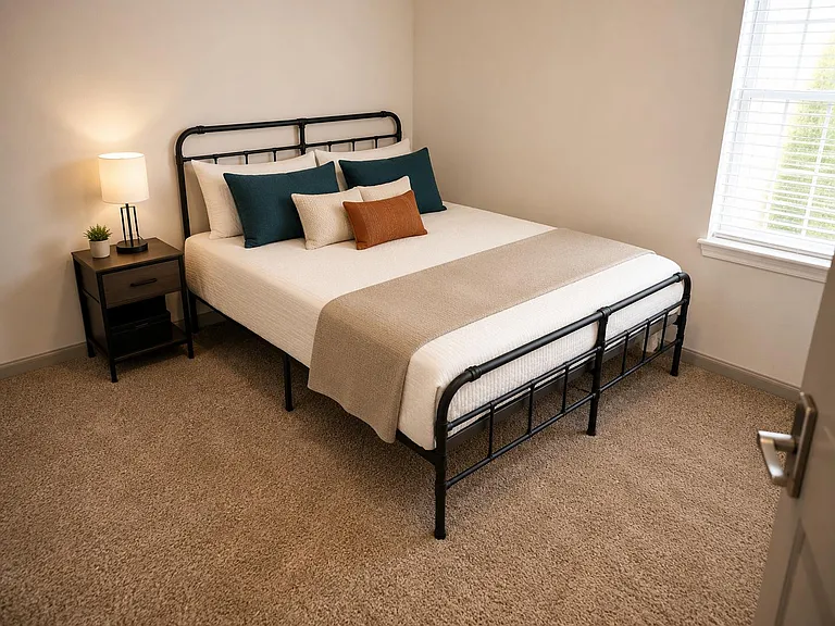 Carpeted bedroom featuring a metal bed frame, nightstand with lamp, and a window with blinds for natural light.