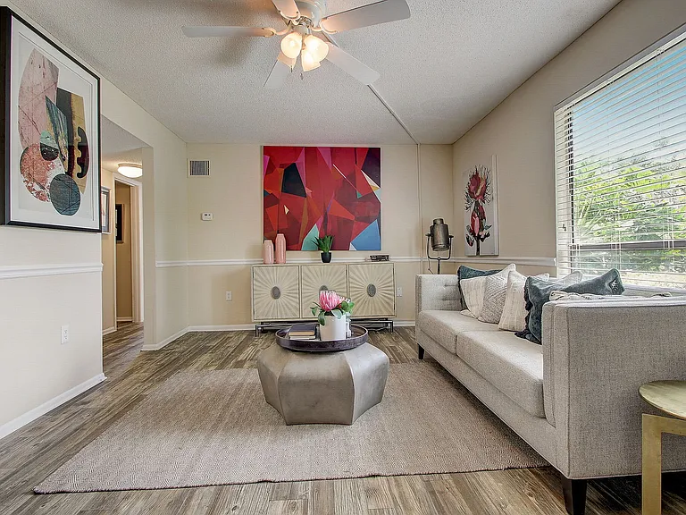 Bright and inviting living room with modern decor, featuring a cozy gray sofa, abstract artwork, and large windows offering lush greenery views.