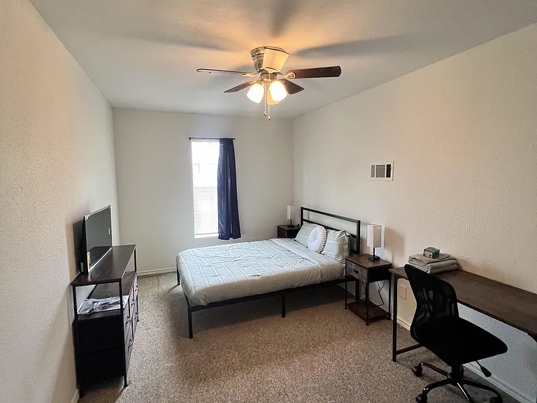 Furnished bedroom with a full bed, nightstands, desk workspace, and a TV stand, plus a window and ceiling fan.