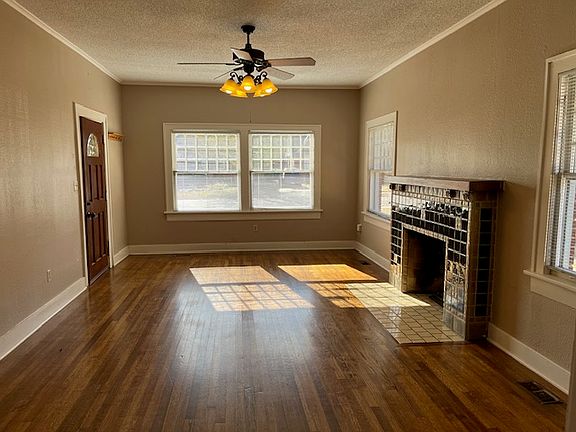 Living room with hardwood flooring, a ceiling fan, a tiled mock fireplace, and plenty of windows allowing natural light.
