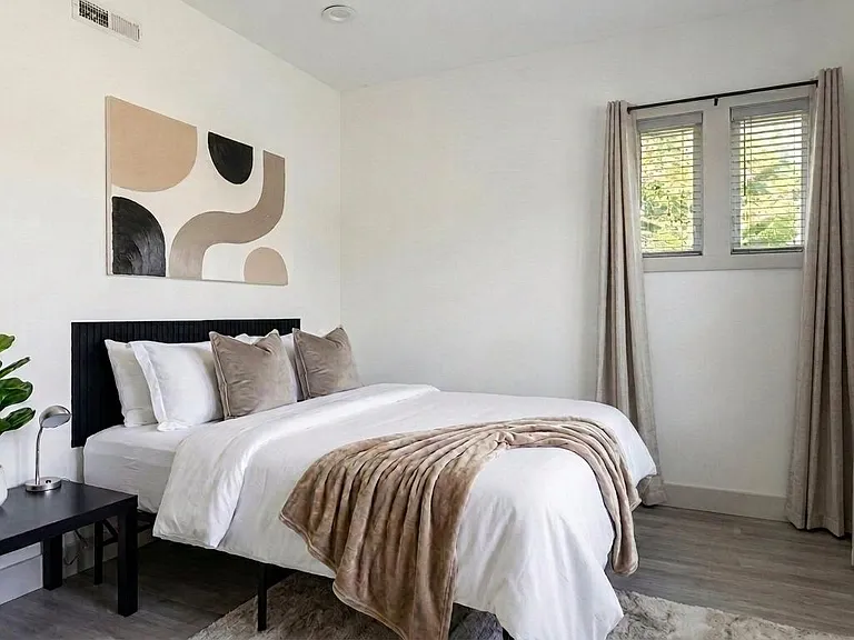 Well-appointed bedroom featuring a queen bed, side table, and large windows with blinds and curtains for light control.
