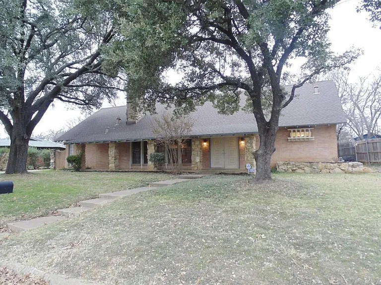 Single-story brick home with a covered front entry, mature shade trees, and a wide front lawn with a walkway to the door.