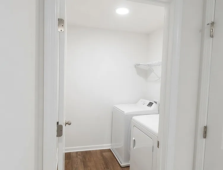 Dedicated laundry room with full-size washer and dryer, upper shelf for storage, and wood-look flooring. Convenient location with good lighting and ventilation.
