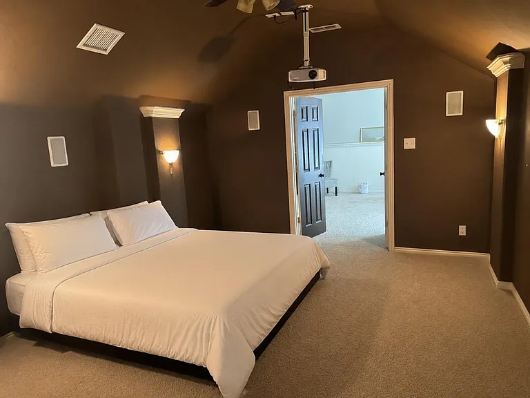 Spacious carpeted bedroom with a comfortable bed, wall sconces, and direct access to an adjacent room or hallway.