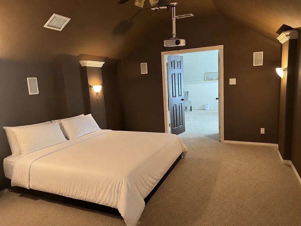 Spacious carpeted bedroom with a comfortable bed, wall sconces, and direct access to an adjacent room or hallway.