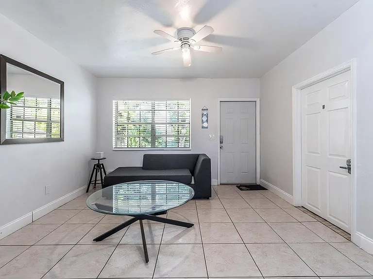 Open living area with tiled floors, ceiling fan, and a large window with blinds for natural light and privacy.