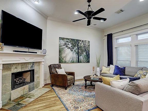 The living room, nestled between oversized windows and a gas-log fireplace with tile surround and a custom mantle, has high ceilings, decorative crown moulding and baseboards, recessed lighting, and gleaming hardwood floors.