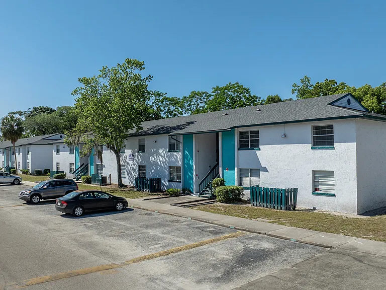 Charming apartment complex with white exteriors and turquoise accents, offering ample parking and lush greenery under a clear blue sky.