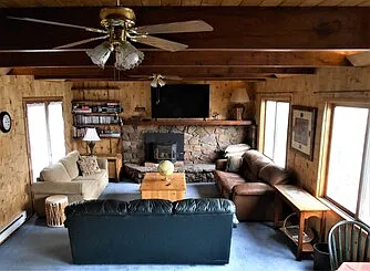 Living room with couches, new 65" smart TV, views of the lake and the deck, beamed ceiling, two ceiling fans, French doors, three big windows, stairs leading up or down, optional desk with file drawers, baseboard heater.