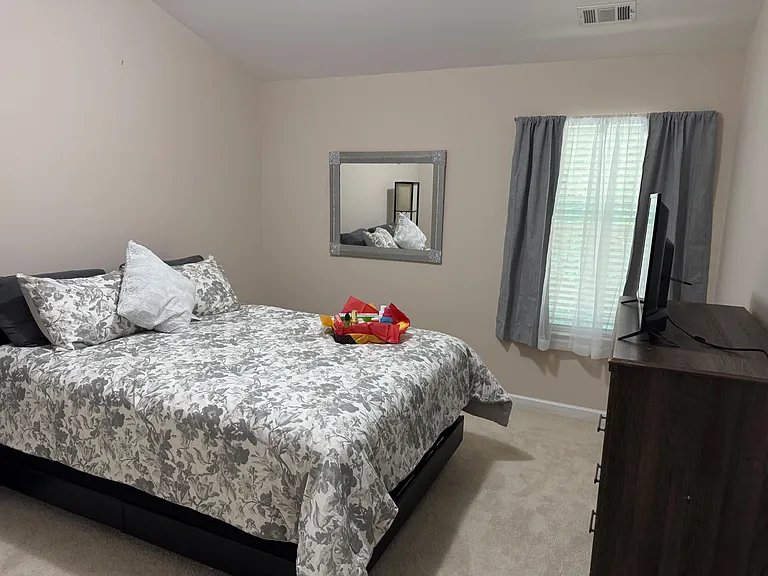 Furnished queens size bedroom, dresser with TV, and a window with blinds and curtains for light and privacy.