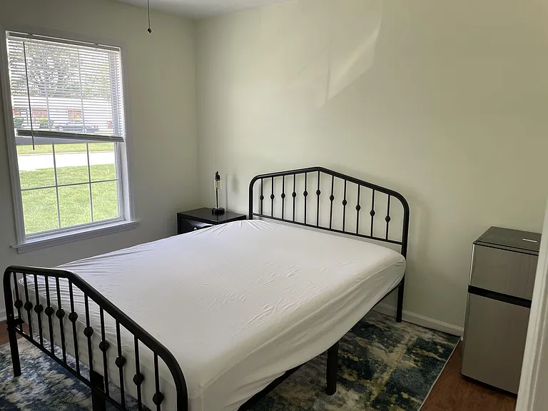Furnished bedroom with a full-size bed, nightstand, mini fridge, and a large window with blinds for natural light.