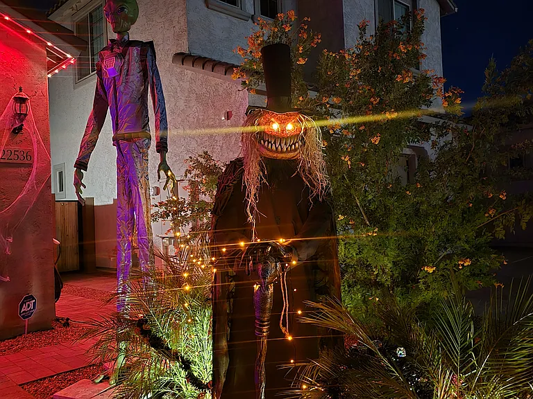 The front yard features towering Halloween animatronics, including a glowing green alien and a separate pumpkin-headed figure. Red and green lights cast an eerie glow, creating a festive, spooky welcome for guests and trick-or-treaters.