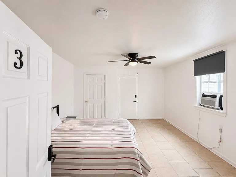 Spacious bedroom with tile flooring, ceiling fan, and a window A/C unit, plus room for a full bed and essentials.
