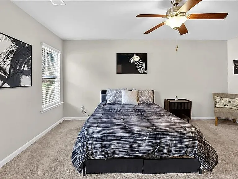 Spacious carpeted bedroom with ceiling fan, large window with blinds, and room for a full or queen bed plus seating.