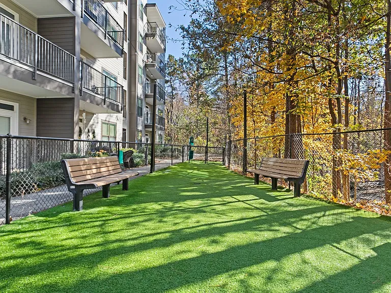 Fenced turf dog park with shaded benches and wooded backdrop to let your pup roam